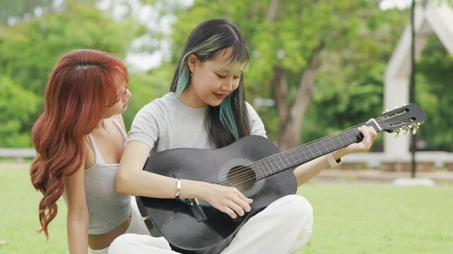 Cheerful Asian lesbian couple playing acoustic guitar and singing together in a sunny green park. Happy young LGBTQ women enjoying romantic musical date and summer freedom.