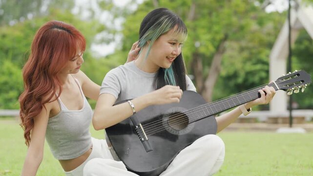 Cheerful Asian lesbian couple playing acoustic guitar and singing together in a sunny green park. Happy young LGBTQ women enjoying romantic musical date and summer freedom.