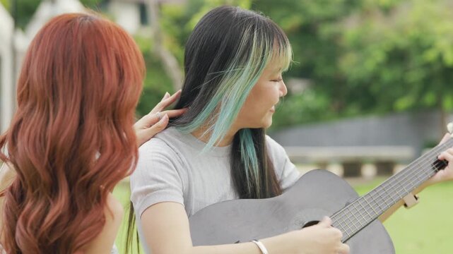 Cheerful Asian lesbian couple playing acoustic guitar and singing together in a sunny green park. Happy young LGBTQ women enjoying romantic musical date and summer freedom.