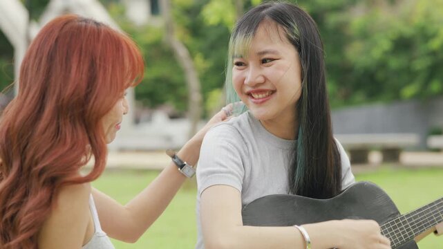 Cheerful Asian lesbian couple playing acoustic guitar and singing together in a sunny green park. Happy young LGBTQ women enjoying romantic musical date and summer freedom.