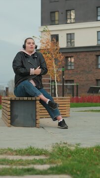 Woman in black leather jacket over pink hoodie rests. Posture is casual legs crossed one ankle over other body. Paved surfaces grassy areas brick structures form backdrop