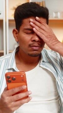 Indian man on home sofa reads bad news on smartphone and feels shock and disappointment during quiet home break. Arabian boy on home sofa reacts to negative message with raised brows and sad stress.