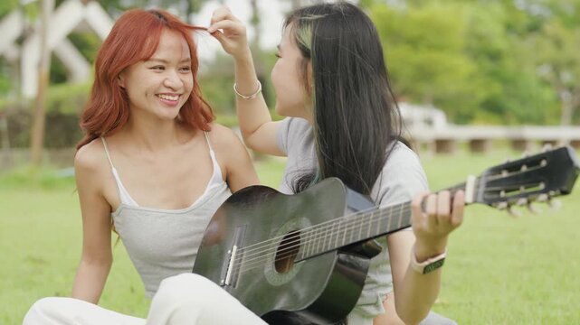 Cheerful Asian lesbian couple playing acoustic guitar and singing together in a sunny green park. Happy young LGBTQ women enjoying romantic musical date and summer freedom.