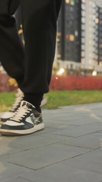 Individual wearing dark trousers with laces and two toned. Secondary individual appears partially visible walking. Outdoor scene features tall structures some illuminated