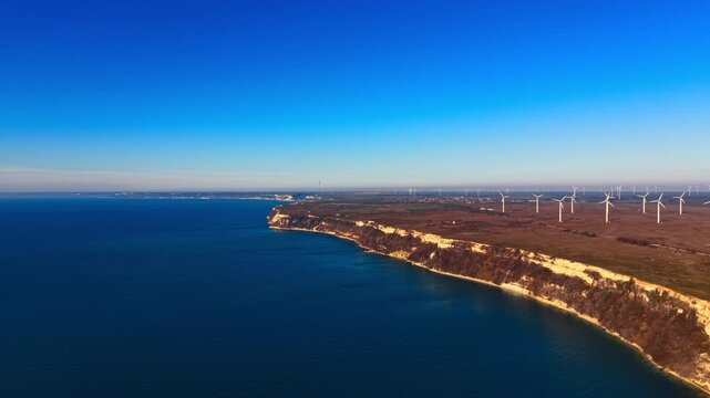 Sunlit turbine rows above chalk cliff coastline, Bulgaria. Elevated view of white towers lined along cliff edge with contrail streaking clear sky.