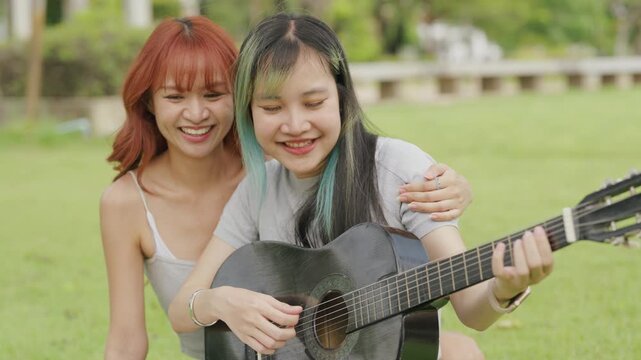 Cheerful Asian lesbian couple playing acoustic guitar and singing together in a sunny green park. Happy young LGBTQ women enjoying romantic musical date and summer freedom.