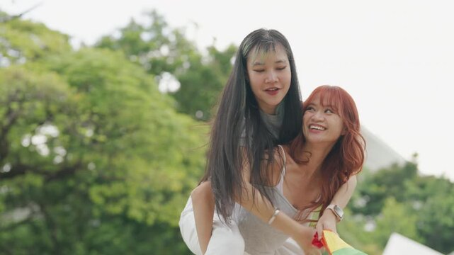 Cheerful Asian lesbian couple playing acoustic guitar and singing together in a sunny green park. Happy young LGBTQ women enjoying romantic musical date and summer freedom.