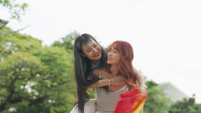 Cheerful Asian lesbian couple playing acoustic guitar and singing together in a sunny green park. Happy young LGBTQ women enjoying romantic musical date and summer freedom.