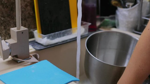 Washing a metal mixer bowl under running water in the kitchen sink with a soapy sponge, cleaning up after homemade baking