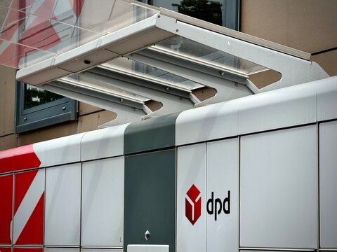 Automated parcel locker station by DPD features a modern design with white and red panels, typical of DPD branding. Located outdoors, it includes a canopy for protection against weather elements.