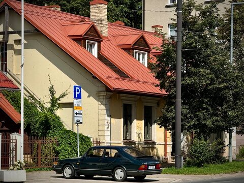 A first-generation Saab 900 hatchback is parked on a street. The car is dark-colored, with a classic curved design typical of the late 20th century. It is parked in front of a yellow house 
