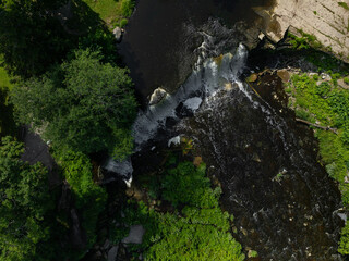 Top down aerial view of Keila-Joa waterfall and Keila River flowing over rocky cascades through dense green forest in northern Estonia. Dark water, white rapids and lush summer vegetation. © Viktoria