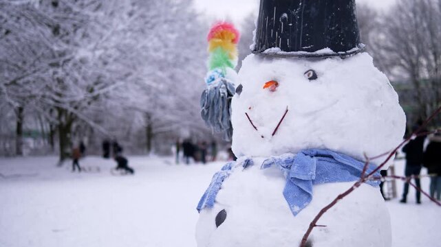 Snowman with a black hat, blue scarf and colorful broom standing in a snowy winter park with people in the background