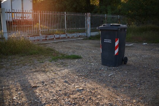 Black plastic garbage bin on gravel near a metal fence and a truck at sunset