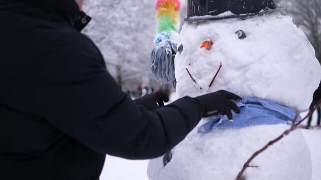 A person wraps a scarf around a snowman during a snowy winter day. The cozy outdoor scene represents winter fun, Christmas holidays, festive traditions