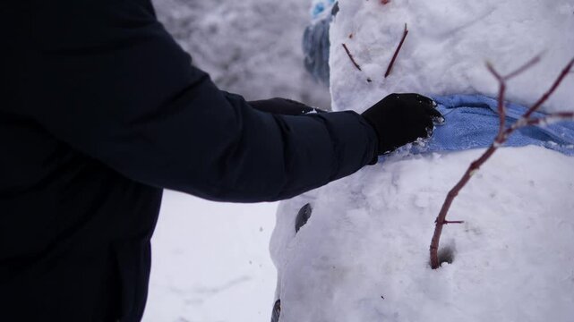 A person puts a warm knitted scarf around a snowman outdoors in fresh winter snow. Cozy scene evoking winter fun, Christmas holidays