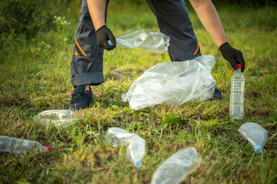 A janitor wearing gloves picks up plastic bottles and wraps them in a bag on the grass. They are participating in a clean-up event to help the environment in a local park