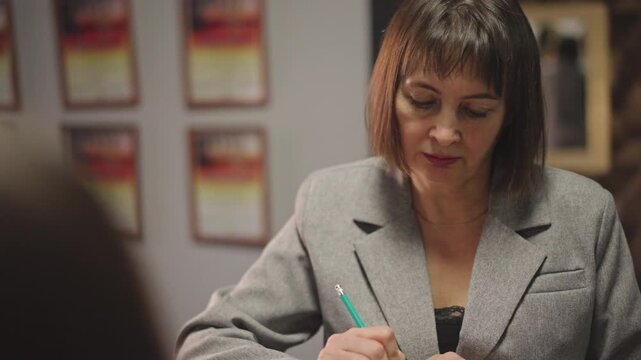 Music school administrator writing enrollment form at reception desk, professional woman signing documents with green pen, framed certificates on wall, warm office lighting, composed administrative