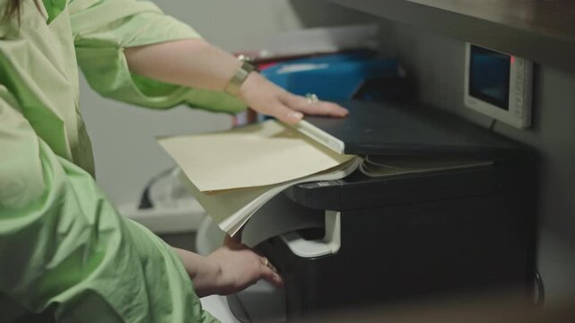 Caucasian music teacher copying sheet music from printer, hands sorting pages in green jacket, organizing stacked scores for school choir rehearsal, closeup on fingers flipping notation, calm focused