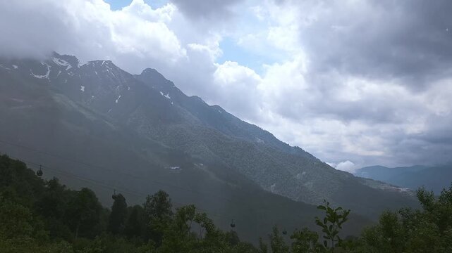 Dark dramatic clouds moving over a majestic mountain range covered with lush green forest, revealing patches of blue sky and snow capped peaks while cable cars are ascending the slope