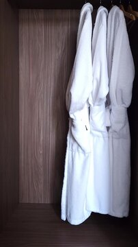 Male hand opening a dark wood wardrobe door with a long metal handle, revealing clean white terry cloth bathrobes hanging inside, a typical amenity in a luxury hotel room or spa