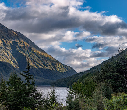 Lake Gunn, Blick durch Rotbuchenwald auf den See mit dramatischem Wolkenhimmel, sonnenbeschienene Bergflanke, Fiordland National Park, Southland, Suedinsel, Neuseeland