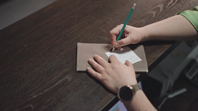 Overhead hands writing on notebook on dark wood desk, smartwatch on wrist, teacher preparing lesson and grading chord diagrams, neat stationery and envelope, focused planning for student practice,