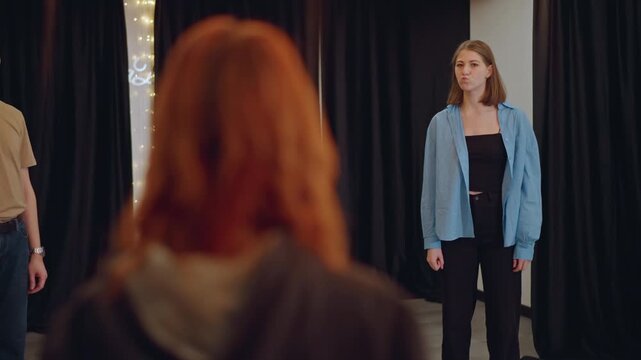 Redheaded coach faces white female singer during audition practice, intimate studio with string lights and black curtain backdrop, student in blue jacket stands center stage while coach watches