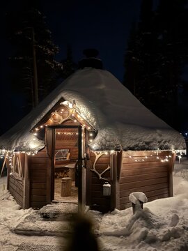 illuminated snow-covered yurt in serene winter forest at night front view with cozy string lights and firewood antlers in warm glow of moonlight and starry sky.