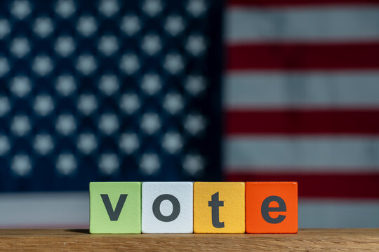 US election voting concept with colorful wooden blocks. The word vote written on colorful wooden blocks on table with blurred American flag in background representing democracy and elections