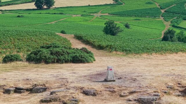 Concrete triangulation pillar marked with red Welsh dragon paint, sunlit dry terrain and green valley pastures during a hot dry spell