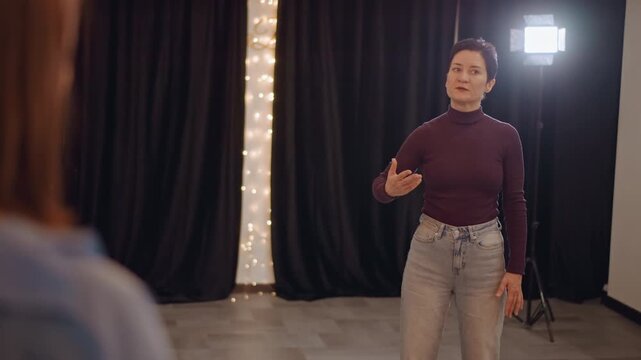 Female performer practices audition monologue under stage lights, receiving director feedback and hand gestures from coach curtain seam and string lights create warm backdrop confident delivery,