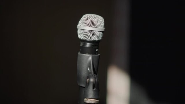 Close up microphone on dark stage, empty rehearsal space with spotlight glow, worn metal mesh and cable, sturdy stand, intimate coaching vibe, gritty texture, low light mood, performer preparation
