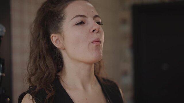 Focused singer practicing breath control and phrasing at microphone, pursed lips and measured exhale during studio lesson, coach evaluates tone and timing, intimate dimly lit rehearsal atmosphere