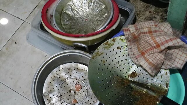 Messy dirty kitchen utensils and metal steamer pots waiting to be washed in the sink.