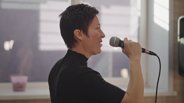 Confident studio performance by white vocalist, standing near window with speaker behind, handheld microphone and bright natural light, amplified sound, expressive delivery, promotional showcase vibe,