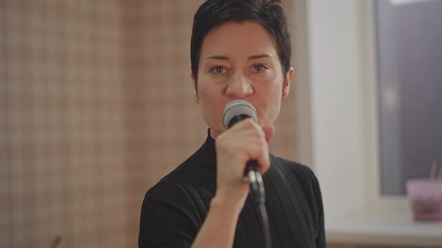Closeup singer delivering vocal lesson in classroom, direct gaze into camera, microphone grip, interpersonal coaching, breath control exercises, muted color palette, window light, attentive posture