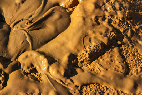 Thick viscous clay sludge covering a muddy ground, wet brown earth texture creating an organic abstract background with smooth surface ripples and drying patches under bright sunlight