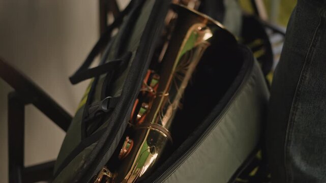 Packing caucasian performer placing sax into padded case after rehearsal or gig, careful hand movements, zipper closing, preparing for travel and commute, evening light and backstage calm.