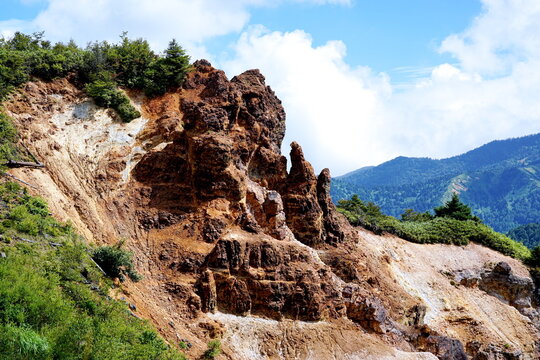 山肌の断崖が露出した自然の景観。岩肌は赤茶色で、緑の植物が点在し、青い空と雲が広がる。
