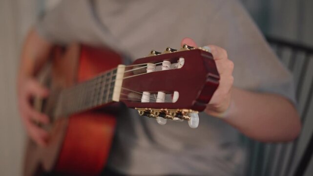 Closeup acoustic guitar headstock and hands tuning during home lesson young student practicing chord progressions on warm wooden guitar, seated on chair in casual room, focused fingers, remote tutor
