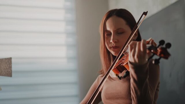 Conscientious violinist concentrates on perfecting performance techniques. Intensely focused musician works on expressive fingerwork during rigorous practice session with natural window light