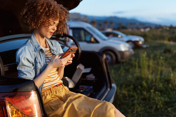 Obraz premium Cheerful woman outdoors, relaxed lifestyle portrait, sitting at car trunk, checking phone, warm sunlight, denim jacket, striped tee, meadow scenery, gentle breeze, natural colors
