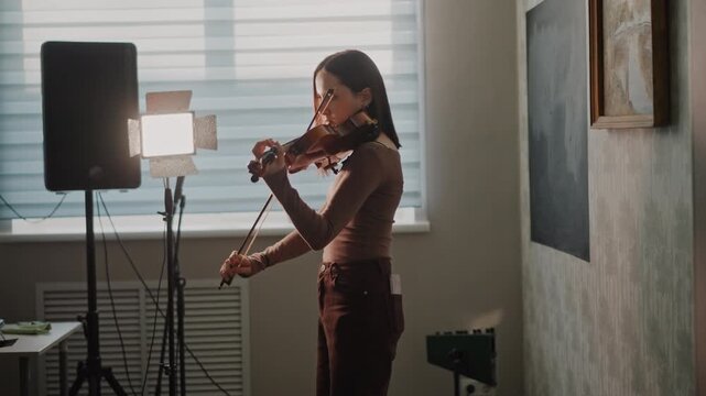White woman rehearsing violin near equipment and speaker, softbox light and blinds, school recital practice turned professional session, focused silhouette and steady bowing, warm ambient tones