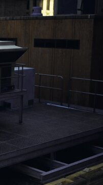 A platform is visible at night in a city. The structure includes railings and a landing area. Nearby, there are station features and walls made of wood. The scene shows urban architecture.