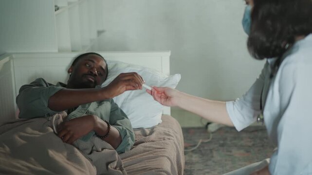 White nurse checking temperature of black patient, thermometer near mouth, vital check and fever concern, bedside interaction, masked clinician recording result, calm clinical mood