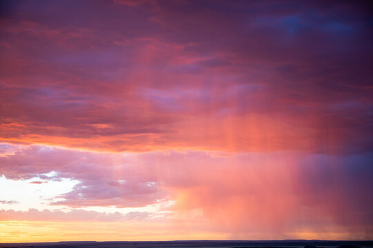 Dramatic dramatic sunset sky featuring vibrant magenta, purple, and orange clouds with a distant rain shaft (virga) illuminated by golden sunlight. High-impact nature background with wide copy space f