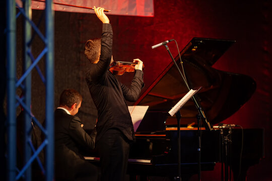 Classical music funding crisis highlights financial pressure on performing artists, with violinist and pianist performing on stage. Funding cuts, cultural policy, budget constraints