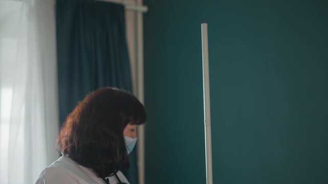 Caucasian doctor adjusts iv pole during home visit wearing surgical mask and gloves, stethoscope around neck, teal curtains and window light in background, preparing infusion tubing and saline bag,