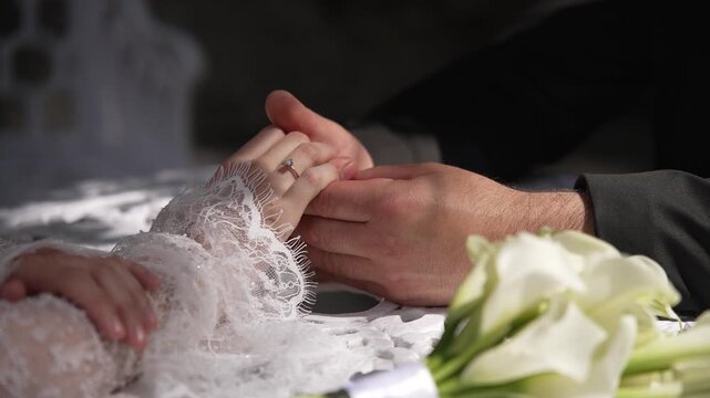 Close-up of a bride and groom holding hands next to a bridal bouquet of white calla lilies, showing an engagement ring.